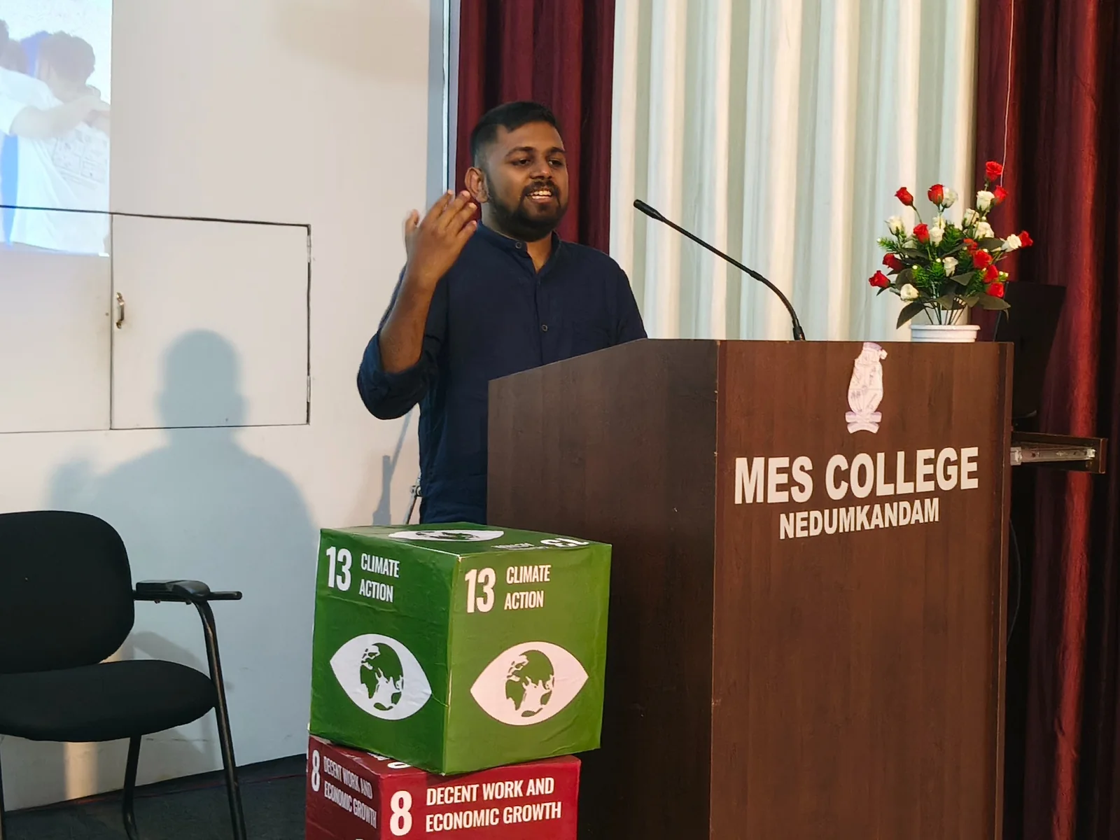 Sanju speaking at a lectern at MES College, Nedumkandam, with Sustainable Development Goal displays nearby